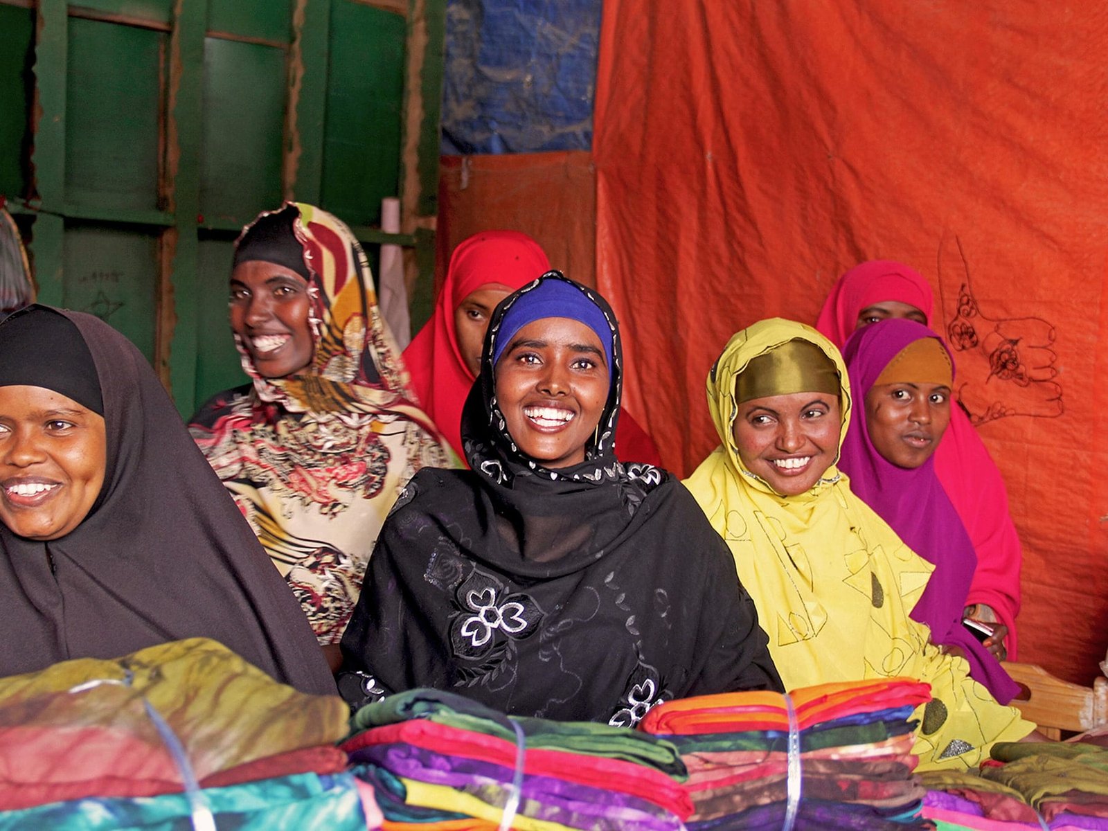 Women seated together behind a market stall stacked with folded, brightly coloured fabrics