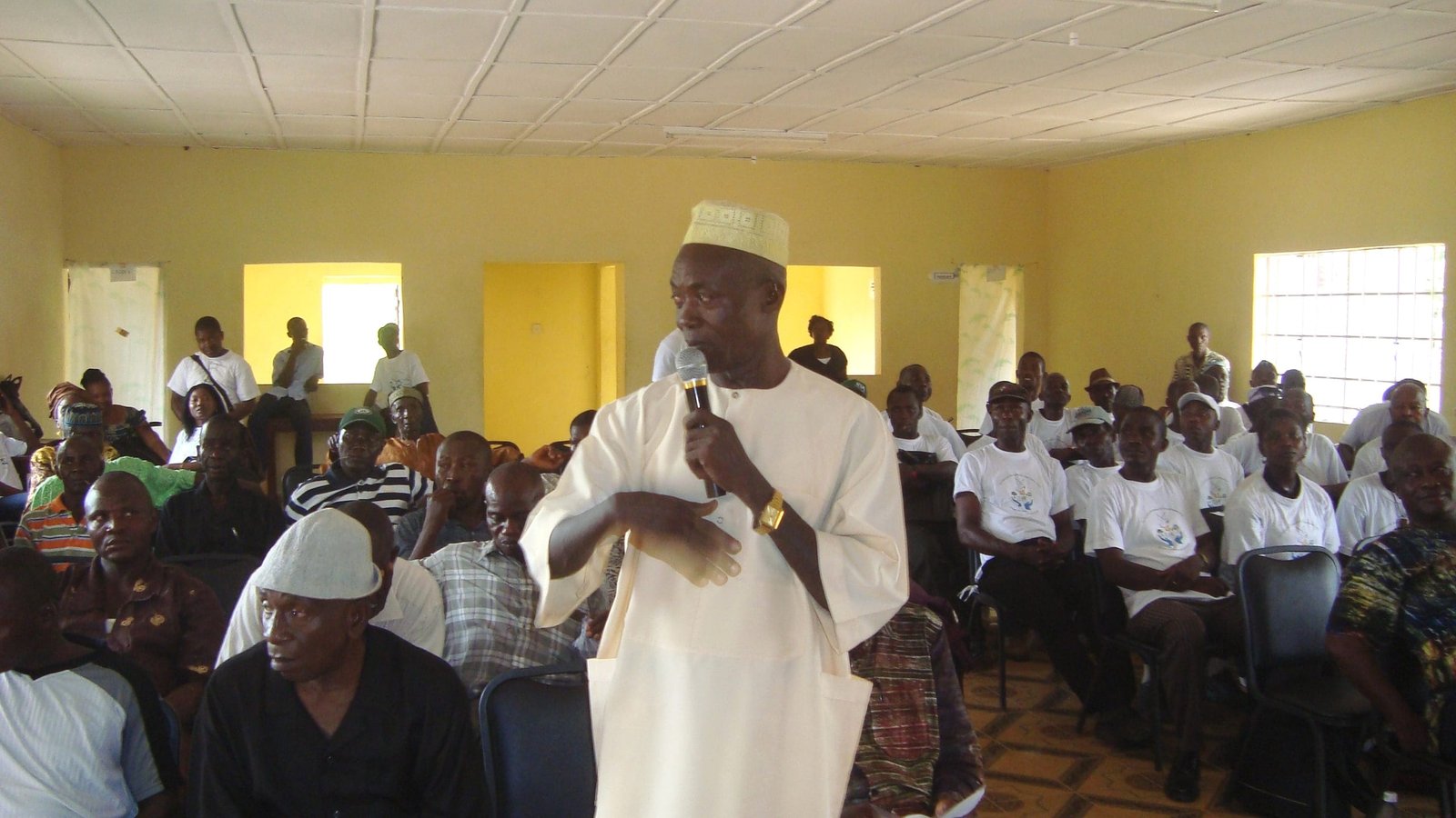 A man speaking into a microphone while addressing a full room at a community meeting